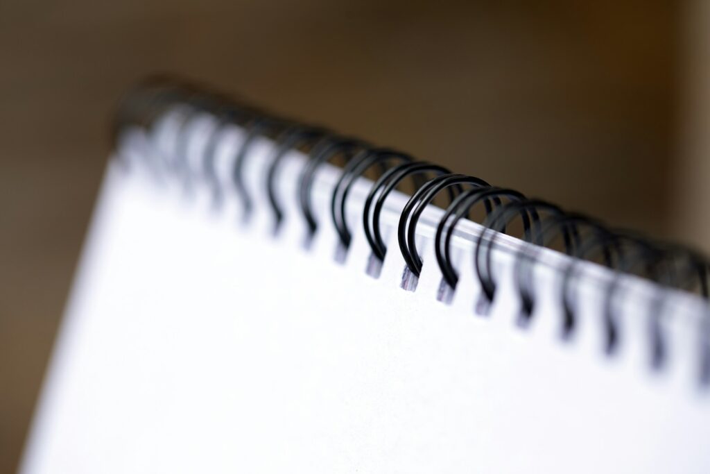 A close-up view of a spiral-bound notebook with black wire binding, focusing on the rings against a blurred neutral background with the use of binding supplies. The pages are white and clean, emphasizing simplicity and functionality.