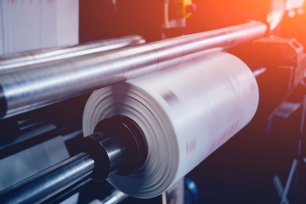 A close-up of a roll of laminating film unwinding on an industrial machine. The metallic rollers and precision mechanism indicate a professional lamination process. A warm glow of light adds a dynamic touch, highlighting the film's smooth texture and the machinery's reflective surfaces.